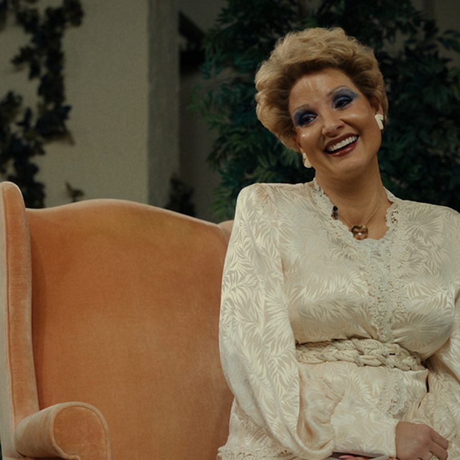 Televangelist Tammy Faye Bakker (Jessica Chastain) sits in an armchair, smiling and wearing heavy makeup, in a scene from The Eyes of Tammy Faye. 